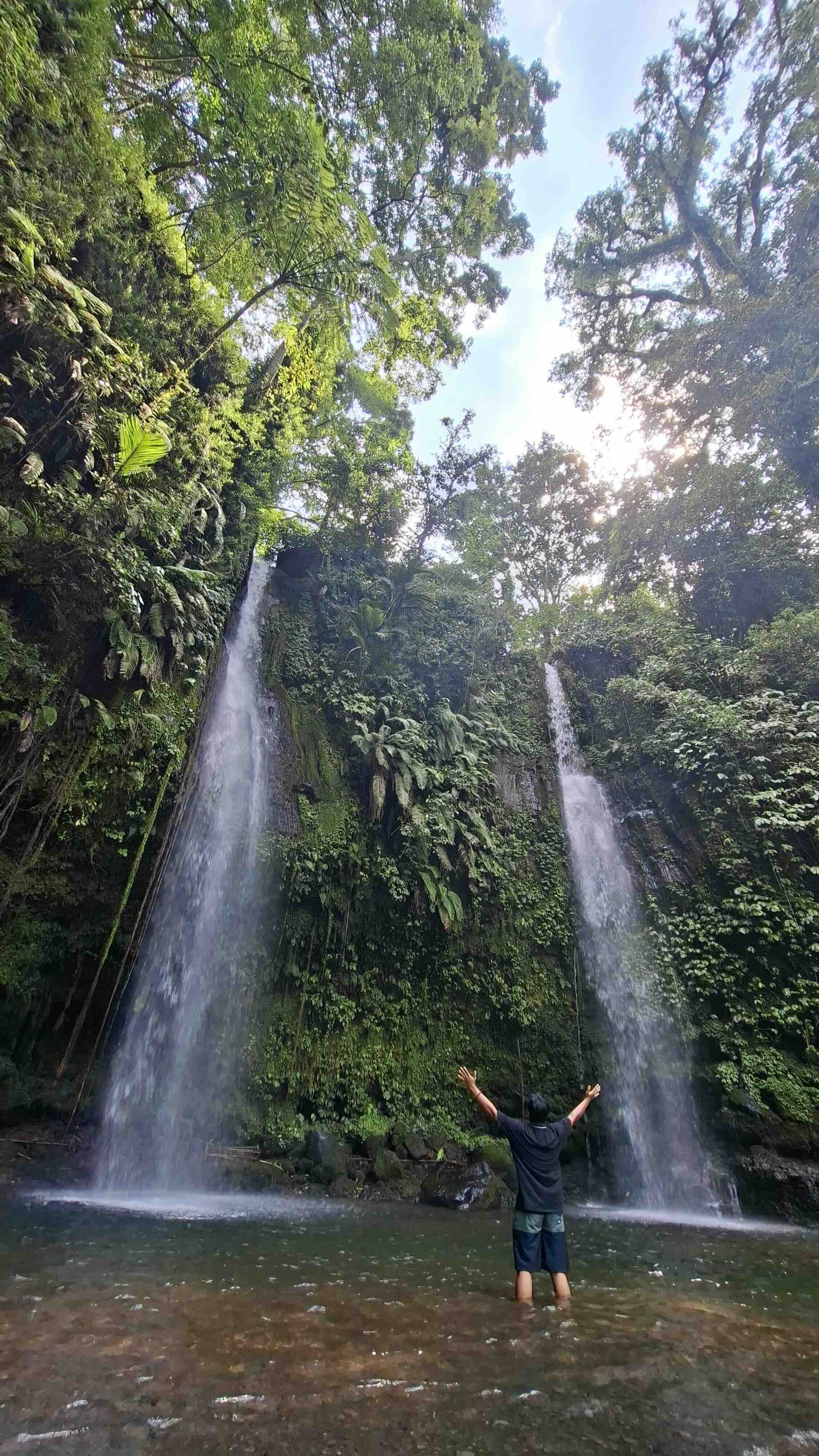 Visitor with arms raised in triumph at twin waterfalls in Tetebatu