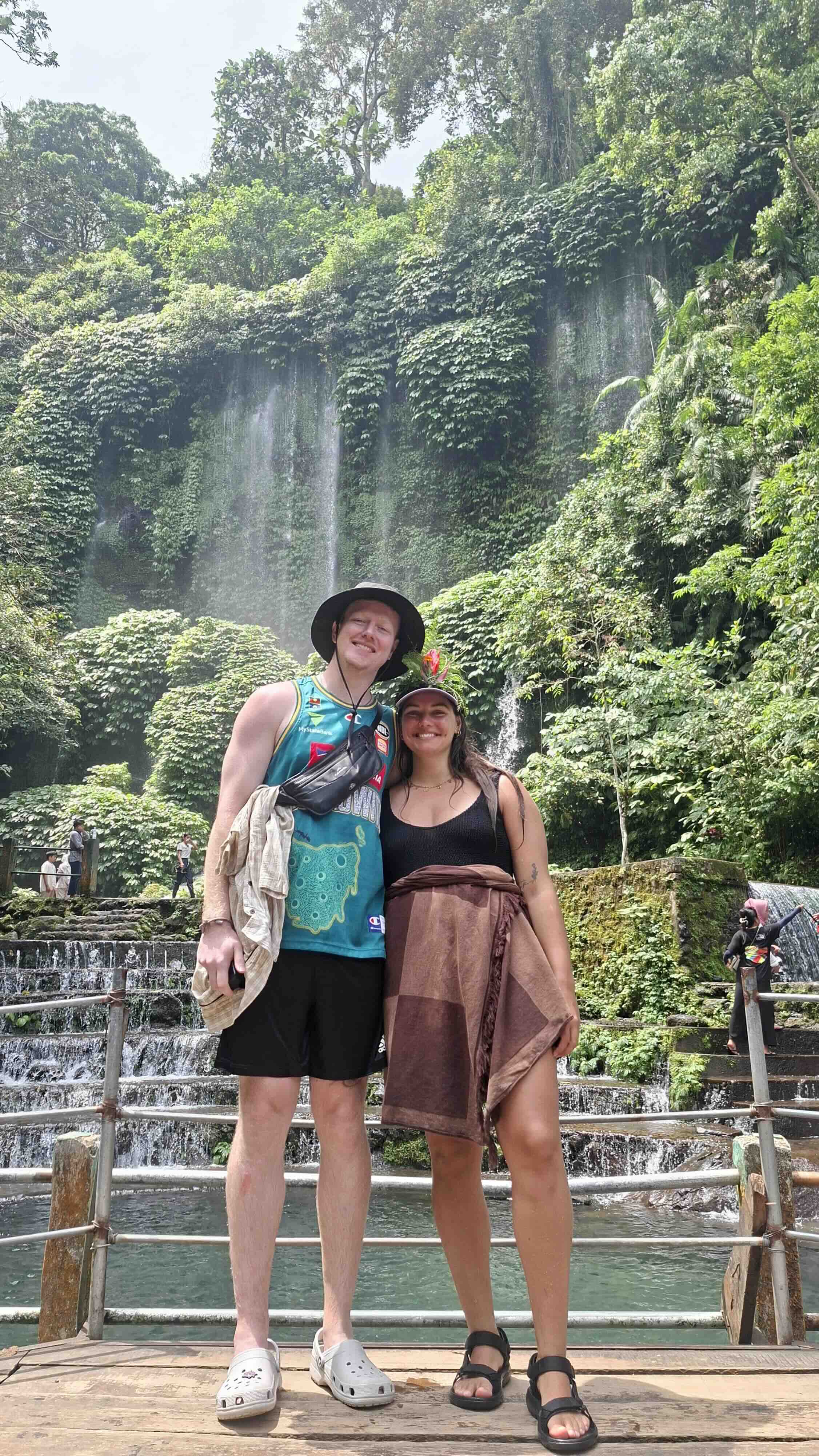 Two hikers standing on a platform with cascading waterfalls behind them in Tetebatu